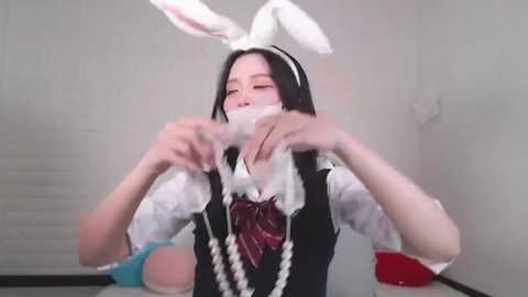 Media: Video of an East Asian woman with pale skin, dressed as a bunny maid, with white bunny ears and a black and white maid outfit. She's using her hands to pull out a fluffy white bunny tail.