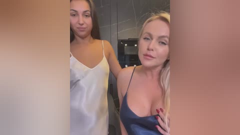 Media: Video of two women in a modern kitchen. The woman on the left has a light complexion, wearing a white, silky camisole. The woman on the right has a fair complexion, large breasts, and is wearing a blue, low-cut camisole. Both have long, straight hair.