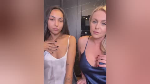 Media: Video of two women with different ethnicities, one with long dark hair in a white camisole, the other blonde in a blue satin camisole, both indoors, in a dimly lit room with a mirror.