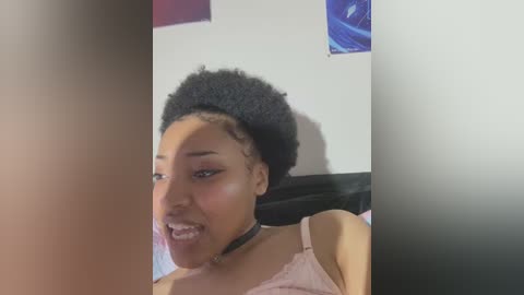 Media: Video of a young Black woman with medium-brown skin, a large afro, and a black choker. She is smiling, wearing a pink top, in a room with blue and purple wall art.