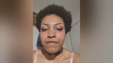 Media: Video of a young Black woman with a medium brown complexion and natural afro hairstyle. She wears pink eyeshadow, a black choker, and a light pink spaghetti strap top. Background is a plain wall with a blue pillow and decorative wall hanging.