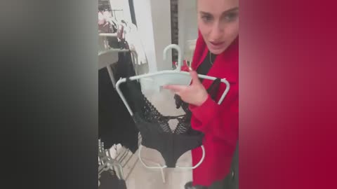 Media: Video of a woman in a red coat holding a black lace bra and panties in a clothing store, with blurred reflections and a red wall in the background.