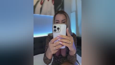 Video of a woman with blonde hair, holding a white iPhone, wearing a leopard print top, sitting on a bed with a black headboard.