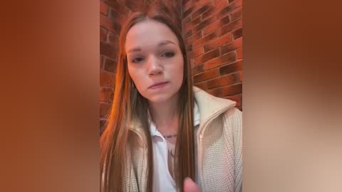 Media: Video of a young Caucasian woman with long, straight blonde hair, wearing a white shirt and beige jacket, standing against a red brick wall, with a warm, soft-focus effect.