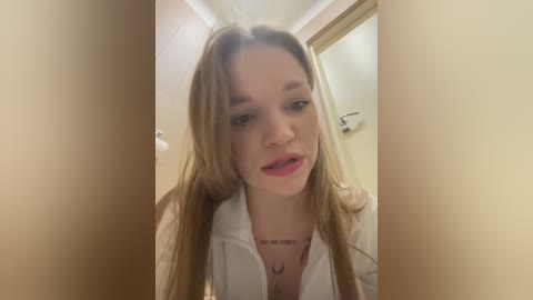 Media: Video of a young woman with long brown hair, wearing a white hoodie, looking directly at the camera. She has light skin and a slight smile. Background shows a bathroom with a door and beige walls.