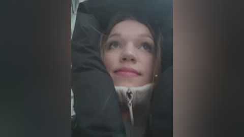 Media: Video of a young woman with fair skin and brown hair, wearing a black jacket, looking up with a thoughtful expression, partially obscured by dark vertical edges.