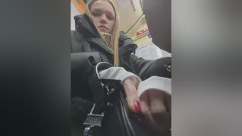 Media: A video shows a blonde woman in a black jacket and white shirt, holding a black handbag, in a dimly lit subway car with blurred passengers and a red sign in the background.