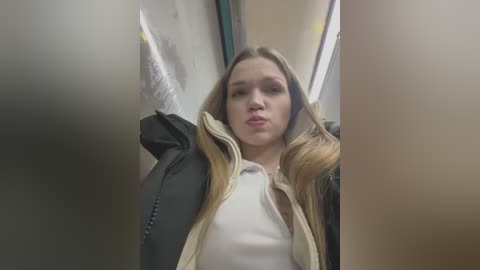 Media: Video of a young Caucasian woman with long, blonde hair, wearing a white shirt under a black, fur-trimmed jacket, looking annoyed in a dimly lit, narrow corridor with beige walls.