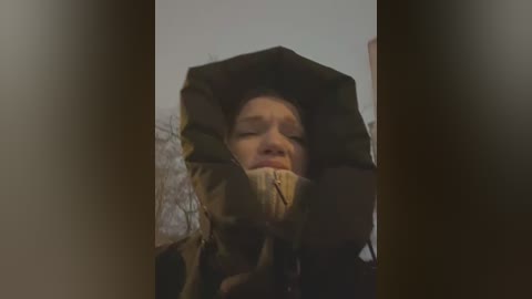 Media: Video of a person with fair skin and short blonde hair wearing a dark hooded jacket, gagged with a brown cloth. Background shows blurred, bare trees.