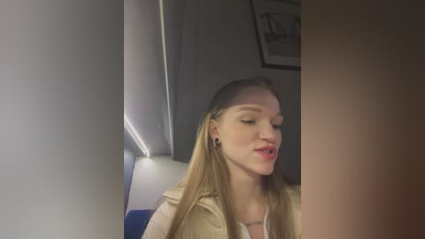 Media: Video of a young, fair-skinned woman with long blonde hair, wearing a beige jacket and a white top, sitting in a plane with dim lighting.