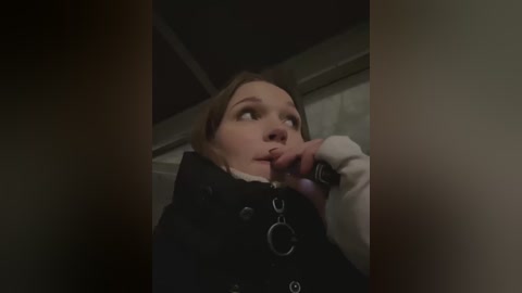 Media: Video of a young Caucasian woman with fair skin and light brown hair, wearing a black coat with metal rings, looking contemplative while holding a phone against her face. Background is dimly lit, suggesting an indoor, possibly urban setting.