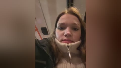 Media: Video of a young woman with light skin, brown hair, wearing a beige turtleneck sweater and black jacket, sitting inside a train, partially obscured by a person in the foreground.