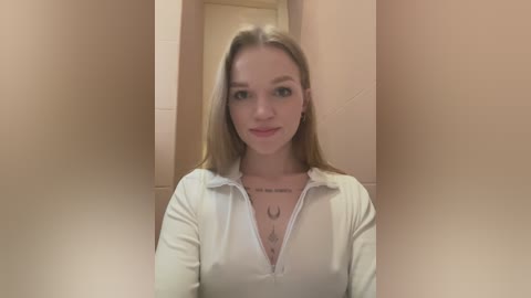 Media: Video of a young Caucasian woman with long blonde hair, wearing a white, unbuttoned blouse, in a beige-tiled bathroom. She has a serene expression and tattoos on her chest.