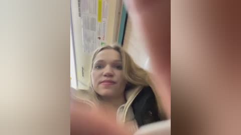 Media: Video of a blonde woman with light skin, wearing a black jacket, sitting in a dimly lit room with posters on the wall.