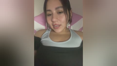 Media: Video of a young woman with light skin and straight, dark hair, wearing a black tank top with a white stripe, sitting on a pink and white striped cushioned chair.