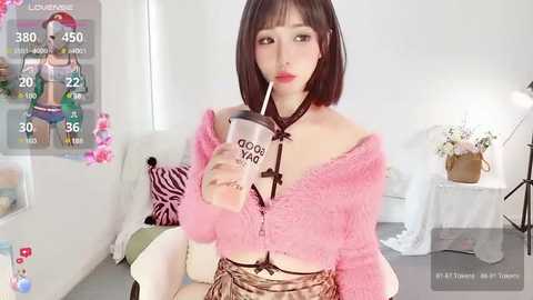 Media: A young Asian woman with straight black hair and a petite frame sits in a cozy living room, sipping from a pink Starbucks cup. She wears a pink fuzzy cardigan and a black bralette, against a background of white walls and floral decor.