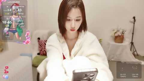 Media: Video of a young Asian woman with shoulder-length brown hair, wearing a white bathrobe, intently using a smartphone. Background features a plain room with minimal decor.