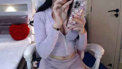 Media: Video of a woman with long black hair, wearing a light purple zip-up hoodie and matching sweatpants, sitting on a white chair in a bedroom. She takes a selfie with a phone case featuring cartoon characters.