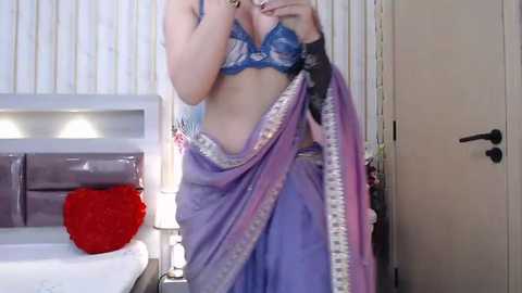 Media: Video of a fair-skinned woman in a blue lace bra, draped in a purple sari with silver trim, standing in a modern bedroom with a white headboard and vertical blinds.