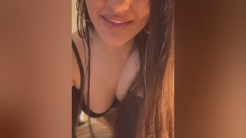 Media: Video of a smiling woman with long dark hair, wearing a black bra, partially visible breasts, in a warm, brown-toned room.