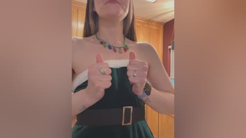 Video of a fair-skinned woman in a green, strapless velvet dress with a Santa Claus belt, wearing colorful beaded jewelry and a ring on her left hand, standing in a warmly lit room with wooden walls and a door.