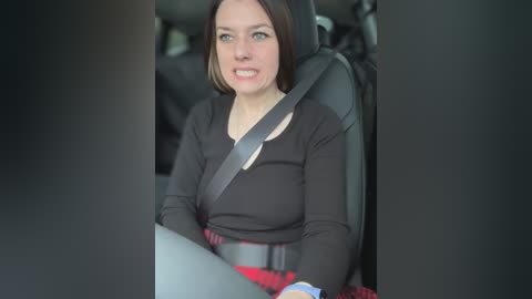 Media: Video of a woman with short brown hair, wearing a black long-sleeved shirt, driving a car with a gray seatbelt across her chest. She looks serious, with a concerned expression. The car's interior is visible in the background.