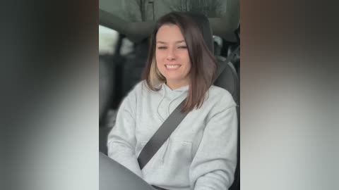 Media: A video of a smiling woman with shoulder-length brown hair, wearing a gray sweater and seatbelt, seated in a car. The background shows the car's interior with a blurred view of the road outside.