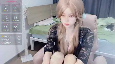 Media: A video of a young woman with fair skin and long blonde hair, wearing a black lace top and a silver necklace, sitting on a beige chair in a messy bedroom.