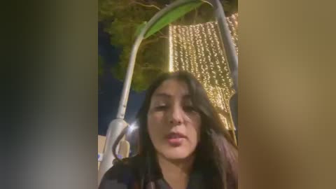 Media: Video of a young woman with medium skin tone, straight black hair, and a neutral expression, sitting outdoors under a streetlamp, with a golden tree in the background, taken at night.