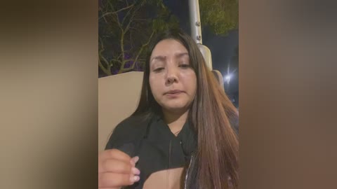 Media: Video of a young Latina woman with long, straight brown hair, wearing a black jacket, sitting in a car at night. She looks somber and is holding her phone.