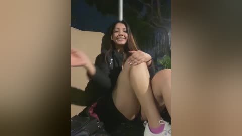 Media: Video of a smiling Latina woman with long dark hair, sitting on a concrete bench at night. She wears a black jacket and shorts, with a purple sock. Background shows blurred trees and a streetlamp.