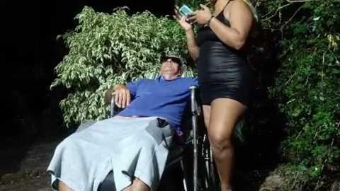 Media: A video captures a man in a wheelchair, wearing a blue shirt and gray pants, surrounded by lush greenery at night. A woman in a black dress stands next to him, holding a phone.