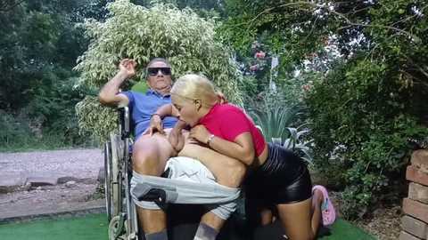 Media: Video of a blonde woman in a red top and black shorts performing oral sex on a bald man in a wheelchair, surrounded by lush greenery.