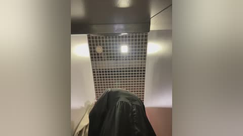 Media: Video of a narrow, dimly lit shower stall with a black ceiling, beige walls, and a square grid of small, square tiles. A towel hangs on the wall.