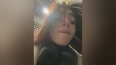 Media: A close-up video of a woman with fair skin, brown hair, and full lips, wearing a black latex bodysuit, standing in a tiled bathroom with a grid-patterned mirror behind her.