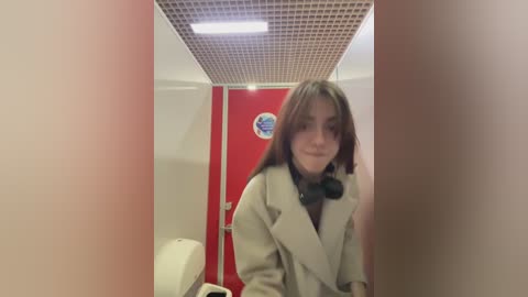 Media: A video of a young woman with long, dark hair and fair skin, wearing a beige coat, standing in a public restroom with a red door and white walls, holding a black headset.