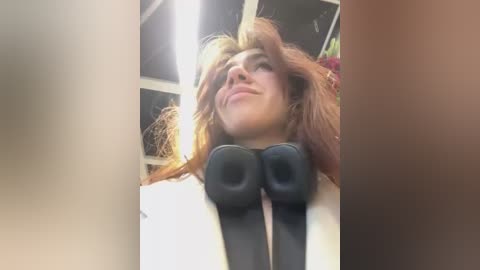 Media: Video of a smiling, light-skinned woman with red hair, wearing a white blazer and black headphones, indoors with a bright ceiling light and blurred background.