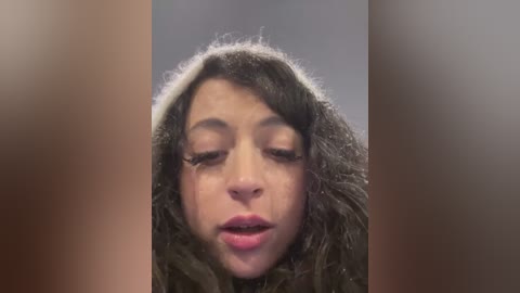 Video of a young woman with curly, shoulder-length brown hair and light skin, her eyes closed, and lips parted, set against a blurred, muted gray background.