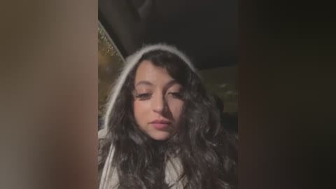 A video of a young woman with curly black hair, wearing a white hooded sweatshirt, against a dimly lit, blurred background.