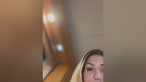 Media: A video of a partially visible woman with light skin and blonde hair, taken in a dimly lit room with blurred wooden furniture and a mirror in the background.