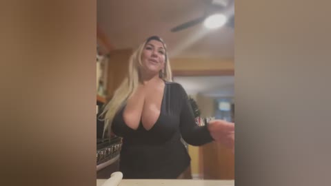 Media: Video of a curvy, blonde woman with large breasts, wearing a black, low-cut top, in a modern kitchen with wooden cabinets and stainless steel appliances.