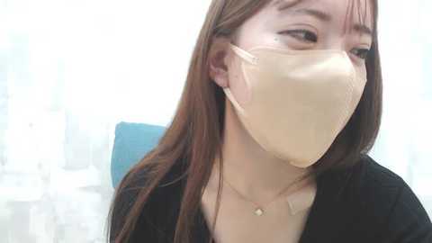 Media: Video of an Asian woman with long brown hair, wearing a beige surgical mask, black top, and a delicate gold necklace. Background shows a blurred blue chair and white curtain.