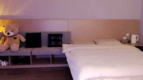 Media: Video of a minimalist hotel room featuring a beige wall, a gray bench with a teddy bear and two pillows, a white bed, and a white nightstand with a white cup.