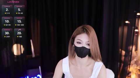 Media: Video of an Asian woman with shoulder-length light brown hair, wearing a white tank top and black face mask, sitting in a dimly lit room with a large screen displaying weather information in the background.