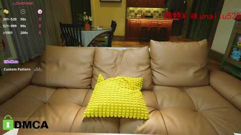 Media: A video of a cozy, dimly-lit living room featuring a beige leather couch with a bright yellow, textured throw pillow. The background reveals a kitchen area with wooden cabinets and a dining table.