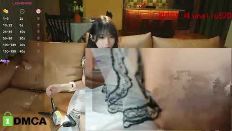 Media: A video captures a young Asian woman with straight black hair, wearing a sleeveless top, sitting on a beige sofa. The background shows a dimly lit room with a kitchen visible. The image is overlaid with a green \"DMCA\" watermark.