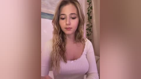 Media: Video of a young Caucasian woman with long, wavy blonde hair, wearing a tight, white ribbed top, standing in a softly lit room with green curtains and a blurred background.