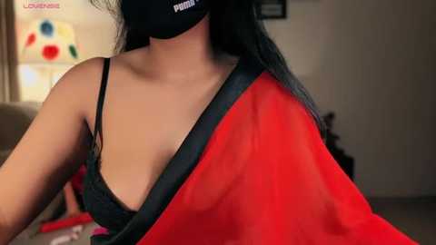 Media: Video of a woman with medium brown skin, wearing a black bra, black mask, and a red sari. She has long black hair, and the background features a blurred living room with a colorful wall art.