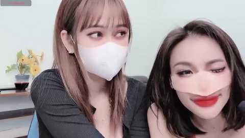 Media: Video of two women with light skin and straight hair, one in a dark top and white mask, the other in red lipstick, smiling. Background shows a white wall with a yellow flower arrangement.