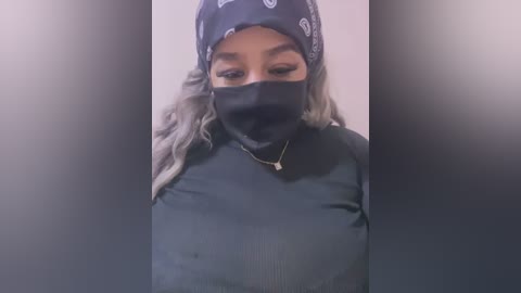 Media: Video of a woman with medium brown skin, wearing a black face mask, long wavy gray hair, and a black bandana. She has a slender figure and is dressed in a black ribbed sweater.